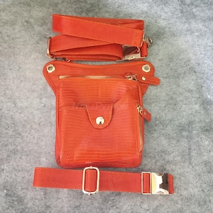 Op de afbeelding: Oranje heuptas met een textuurpatroon dat op reptielhuid lijkt. De tas heeft een voorvak met een gouden knoop, een hoofdvak met ritssluiting en een verstelbare riem met een gouden gesp. Er is ook een schouderriem. Afmetingen: ongeveer 25 cm x 15 cm.