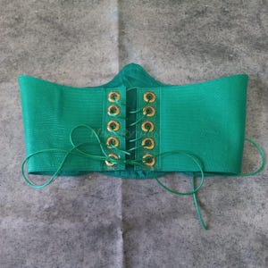 Könnte beinhalten: Ein leuchtend grüner Korsettgürtel mit einem strukturierten, reptilienartigen Muster. Der Gürtel hat goldfarbene Ösen und einen passenden grünen Schnürverschluss. Das Korsett ist so konzipiert, dass es die Taille betont und jedem Outfit einen stilvollen Akzent verleiht.