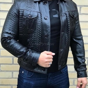 Pode incluir: Uma jaqueta de couro preta com um padr&atilde;o de pele de cobra. A jaqueta tem dois bolsos no peito, bot&otilde;es e gola. A pessoa est&aacute; vestindo uma camiseta preta e jeans azul.