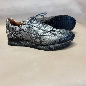 Pode incluir: Um par de tênis cinza e preto com padrão de pele de cobra. Os tênis têm uma sola de borracha branca e um fecho de cadarço preto.