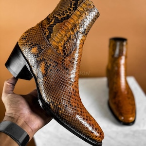 Op de afbeelding: Een paar bruine leren laarzen met een slangenhuidpatroon. De laarzen hebben een spitse neus en een blokhak.