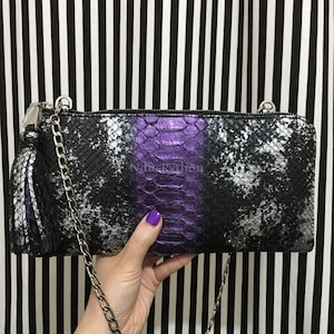 Peut inclure: Un sac à main rectangulaire noir et argenté à motif peau de serpent, avec un panneau central violet. Le sac est doté d'une bandoulière en chaîne argentée et d'un pompon assorti. Le fond est à rayures noires et blanches.
