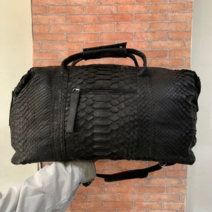 May include: A black leather duffel bag with a snakeskin pattern. The bag has a top handle and a shoulder strap.