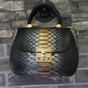 Peut inclure: Un sac à main noir et doré avec un motif peau de serpent. Le sac est doté d'une poignée supérieure et d'une fermeture à fermoir doré. Il est suspendu à un crochet noir sur un mur de briques grises.