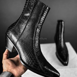 Peut inclure: Bottine noire en cuir avec un bout pointu et un talon épais. La botte présente un motif texturé semblable à de la peau de serpent et une fermeture éclair latérale. Une autre botte est visible en arrière-plan.