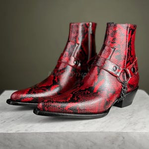 Op de afbeelding: Een paar rode cowboylaarzen van slangenleer met een spitse neus, een gespsluiting en een kettingdetail.