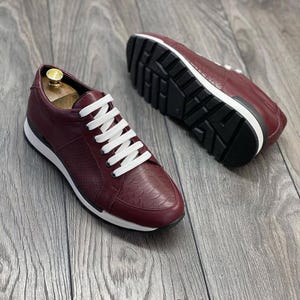 Pode incluir: Um par de tênis de couro bordô com cadarços brancos. Os tênis têm uma parte superior de couro texturizado e uma sola de borracha branca.