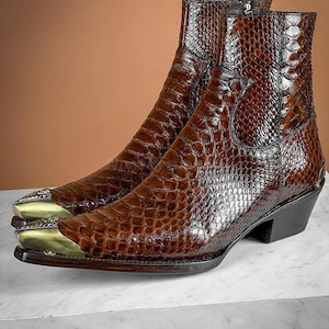 Könnte beinhalten: Ein Paar braune Cowboy-Stiefel aus Leder mit Schlangenhautmuster und goldenen Metallverzierungen an den Zehen.