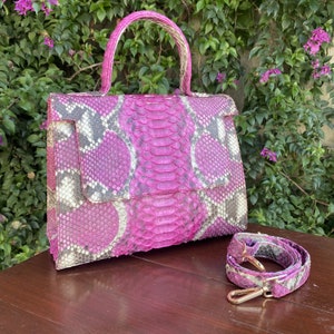 May include: A pink and gray snakeskin handbag with a matching shoulder strap. The bag has a top handle and a flap closure. The strap has gold-colored hardware. The bag is sitting on a brown surface.