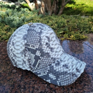 Può includere: Cappellino da baseball con motivo a pelle di serpente nei toni del grigio e del bianco. Il cappellino ha una visiera curva e un bottone sulla parte superiore. Il cappello è realizzato in un materiale strutturato, su sfondo verde.