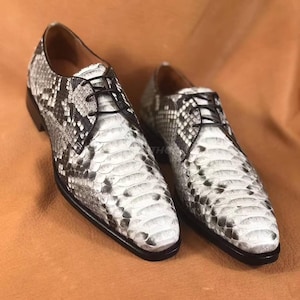 May include: A pair of white and grey snakeskin leather dress shoes with black laces. The shoes have a pointed toe and a low heel.