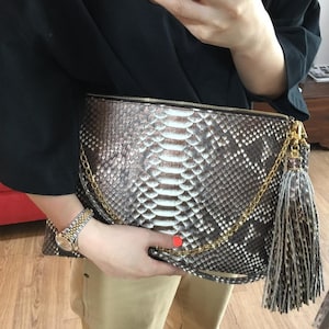 Puede incluir: Un clutch rectangular con un estampado de piel de serpiente en tonos marrón, blanco y gris. Cuenta con una cremallera dorada, una cadena y una borla. El clutch está siendo sostenido por una persona.