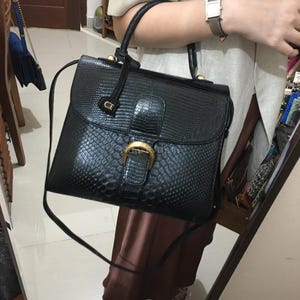 May include: A white ceramic mug with a black handle and the text "But First, Coffee" in a bold, black font. The mug is set against a blurred background, suggesting a cozy setting.