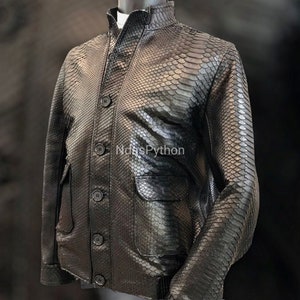 Op de afbeelding: Een zwart leren jack met een slangenhuidpatroon. Het jack heeft een knoopsluiting en een klepzak aan de linkerkant.