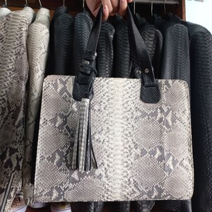 Może przedstawiać: Prostokątna torebka ze wzorem skóry węża w odcieniach szarości i bieli, z czarnymi skórzanymi uchwytami i frędzlami. Torebka jest prezentowana przed wieszakiem z skórzanymi kurtkami.