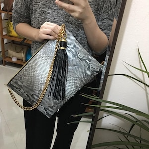 Puede incluir: Bolso de mano con estampado de piel de serpiente gris y blanco, con cadena dorada y borla negra. El bolso es rectangular con cierre de cremallera. La cadena dorada mide unos 100 cm.