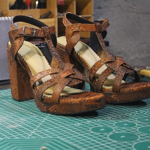 Puede incluir: Un par de sandalias de tacón de cuero marrón con un diseño de tiras cruzadas. Las sandalias tienen un tacón grueso y están hechas de cuero texturizado.