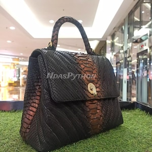 May include: A black and brown snakeskin handbag with a top handle. The bag has a flap closure with a gold clasp. The bag is made of genuine python skin.
