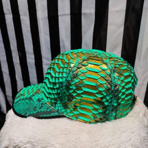 Peut inclure: Une casquette de baseball en peau de serpent verte et dorée. La casquette a une visière incurvée et une couronne structurée.