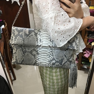 Puede incluir: Un clutch con estampado de piel de serpiente en tonos grises y blancos, con una cadena plateada y una borla a juego. El bolso rectangular parece ser de cuero. El clutch está doblado en la parte superior.