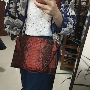 May include: A red and black python skin clutch with a gold chain strap. The bag has a rectangular shape and a tassel detail. The text "NdasPython" is visible on the bag.