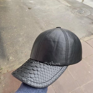 Może przedstawiać: Czarna skórzana czapka bejsbolówka z daszkiem w wzór skóry węża. Czapka ma wszytą szw w środku korony.