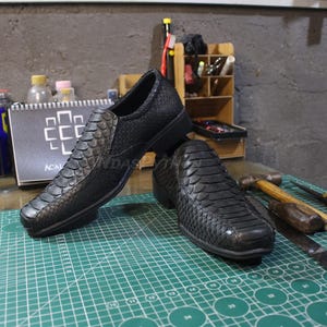 Puede incluir: Dos zapatos de cuero negro con estampado de piel de serpiente. Los zapatos tienen una punta puntiaguda y un tacón bajo. Los zapatos están sobre una estera de corte verde.