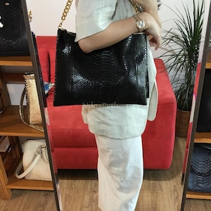 Puede incluir: Un bolso tote negro con un estampado de piel de serpiente y una correa de cadena dorada. El bolso lo sostiene una persona frente a un espejo. El bolso tiene forma rectangular y cierre de cremallera.