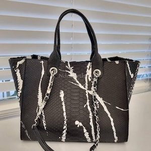 Op de afbeelding: Zwarte en witte tote bag met een slangenpatroon en witte verfachtige strepen. De tas heeft twee zwarte handvatten en een zilveren kettingriem. De tas staat op een wit oppervlak.