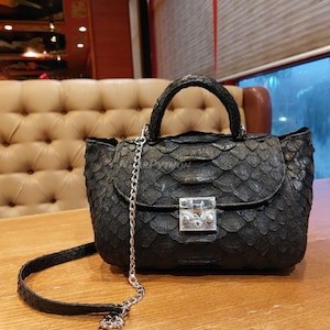 May include: A black leather handbag with a silver chain strap and a silver lock closure. The bag has a textured, scaly pattern, reminiscent of snakeskin.