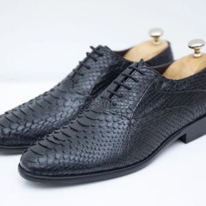 Pode incluir: Um par de sapatos de couro preto com padrão de pele de cobra. Os sapatos têm fecho de atacadores e bico pontudo.