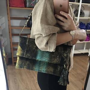Puede incluir: Un clutch negro y dorado con un centro turquesa, con un patrón de piel de serpiente. El bolso tiene una correa de cadena dorada y una borla negra. El bolso está siendo sostenido por una persona.