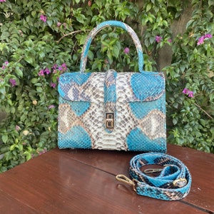 Puede incluir: Un bolso de mano de piel de serpiente con asa superior y bandolera extraíble. El bolso presenta un estampado en tonos turquesa, blanco y marrón con un cierre dorado. La correa y el asa coinciden con el estampado del bolso.