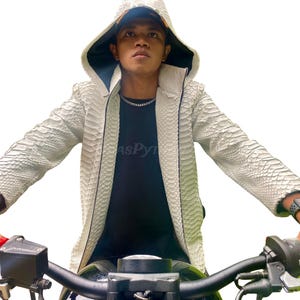 May include: A white snakeskin-patterned jacket with a hood and zip closure. The jacket has a dark lining. The person in the image is wearing a black t-shirt and a silver chain. The jacket is the main focus of the image.