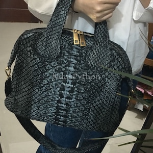 May include: A black and grey snakeskin handbag with gold hardware and a long strap. The bag has a top zipper closure and a structured shape.