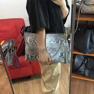 May include: A snakeskin-patterned clutch with a silver chain strap and a tassel. The bag is gray and white with a zipper closure. Other bags are visible in the background.