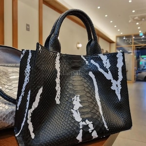 Könnte beinhalten: Schwarze Tragetasche mit weißem, abstraktem Muster. Die Tasche hat eine strukturierte Oberfläche und zwei kurze Griffe. Der Text "dasPython" ist auf der Tasche zu sehen. Die Tasche besteht aus einem Material, das Schlangenhaut ähnelt.