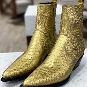 Puede incluir: Un par de botas vaqueras doradas con punta puntiaguda y estampado de piel de serpiente. Las botas son de cuero y tienen un tacón negro.