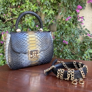 May include: A black and gold snakeskin-patterned handbag with a top handle and gold hardware. A matching black snakeskin bracelet with gold chain links and two gold rings are also present.