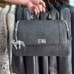 May include: A black leather handbag with a silver clasp and a silver horse charm. The bag is made of snakeskin and has a textured surface.