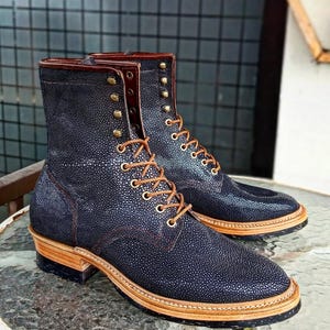 Puede incluir: Un par de botas de cuero azul oscuro con cordones marrones y una superficie texturizada. Las botas tienen una suela gruesa y un ribete de cuero marrón alrededor de la parte superior.