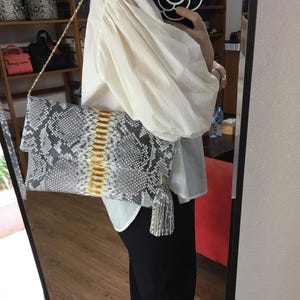 Peut inclure: Une pochette à motif peau de serpent gris et blanc avec une bandoulière chaîne dorée et une bande centrale dorée et jaune. La pochette a un pompon assorti et est tenue devant une personne.