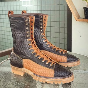 Puede incluir: Un par de botas de cuero marrón y negro con un patrón de piel de serpiente. Las botas tienen un cierre de encaje y una suela de goma gruesa.