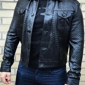 Puede incluir: Chaqueta de cuero negra con un patrón de piel de serpiente, con cierre de botones, dos bolsillos en el pecho y cuello. La chaqueta se lleva sobre una camiseta negra y combinada con vaqueros azules.