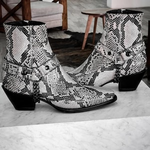 Puede incluir: Un par de botas vaqueras de piel de serpiente negra y blanca con un detalle de cadena y una punta puntiaguda.