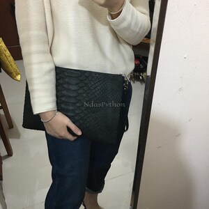 May include: A black clutch with a textured snakeskin pattern. The rectangular bag has a zipper closure and a wrist strap. The text "NdasPython" is visible on the front of the bag. The bag is held in front of a person wearing a white sweater and blue jeans.