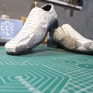May include: Two white leather shoes with a snakeskin pattern. The shoes have a black sole and are on a green cutting mat.