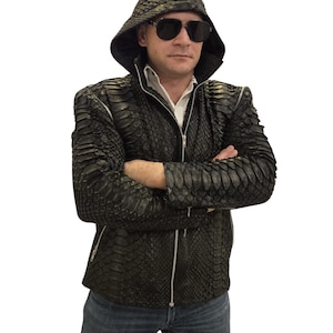 Puede incluir: Chaqueta negra con capucha y estampado de piel de serpiente. La chaqueta tiene cierre de cremallera y una camisa blanca con cuello debajo. La persona lleva gafas de sol y vaqueros azules.