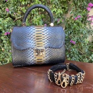 May include: A black and gold snakeskin-patterned handbag with a top handle and gold hardware, paired with a matching black bracelet featuring gold chain detailing and clasps. The bag has a rectangular shape.