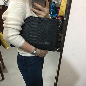May include: Black clutch with a snakeskin pattern. The clutch has a zipper closure and a wrist strap. The text "NdasPython" is visible on the front of the clutch. The clutch is held by a person wearing a white sweater and blue jeans.